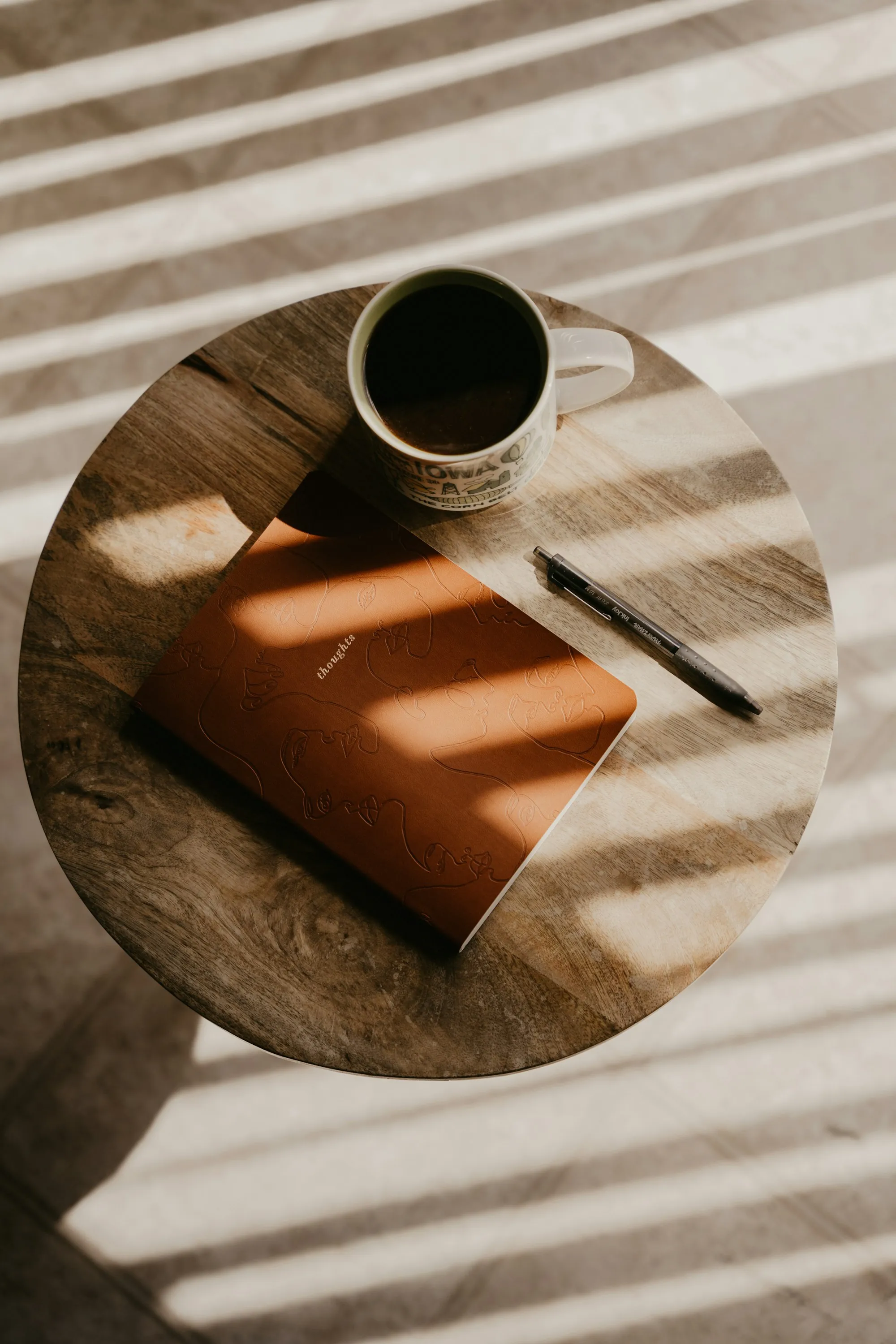 A journal and a cup of coffee on a round wooden table, striped sunlight falling across them